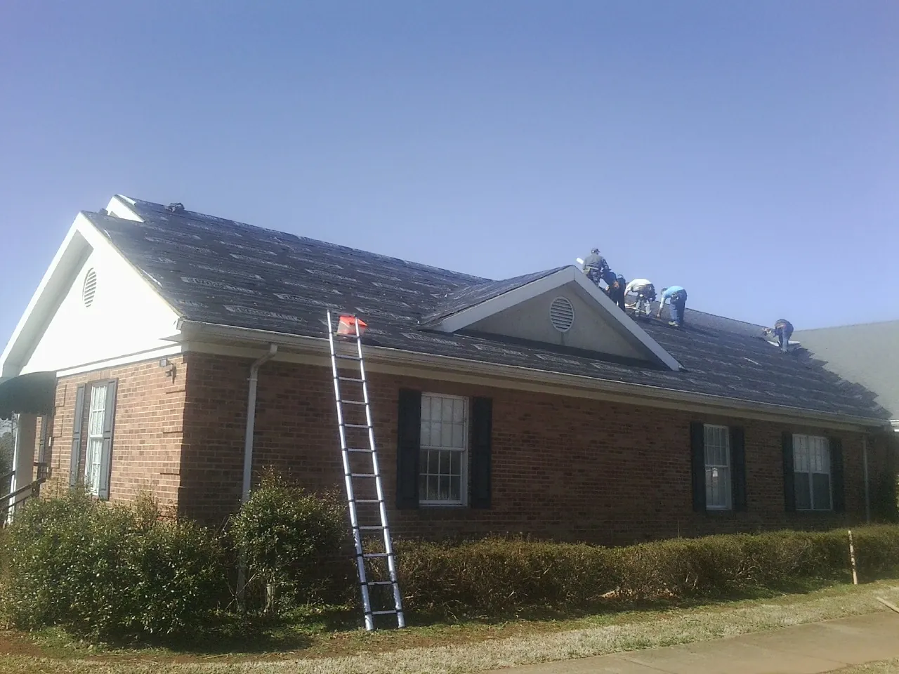 Crew tearing shingles off a brick ranch roof with a ladder set — Metal Roof Repair work by our crew in Bedford