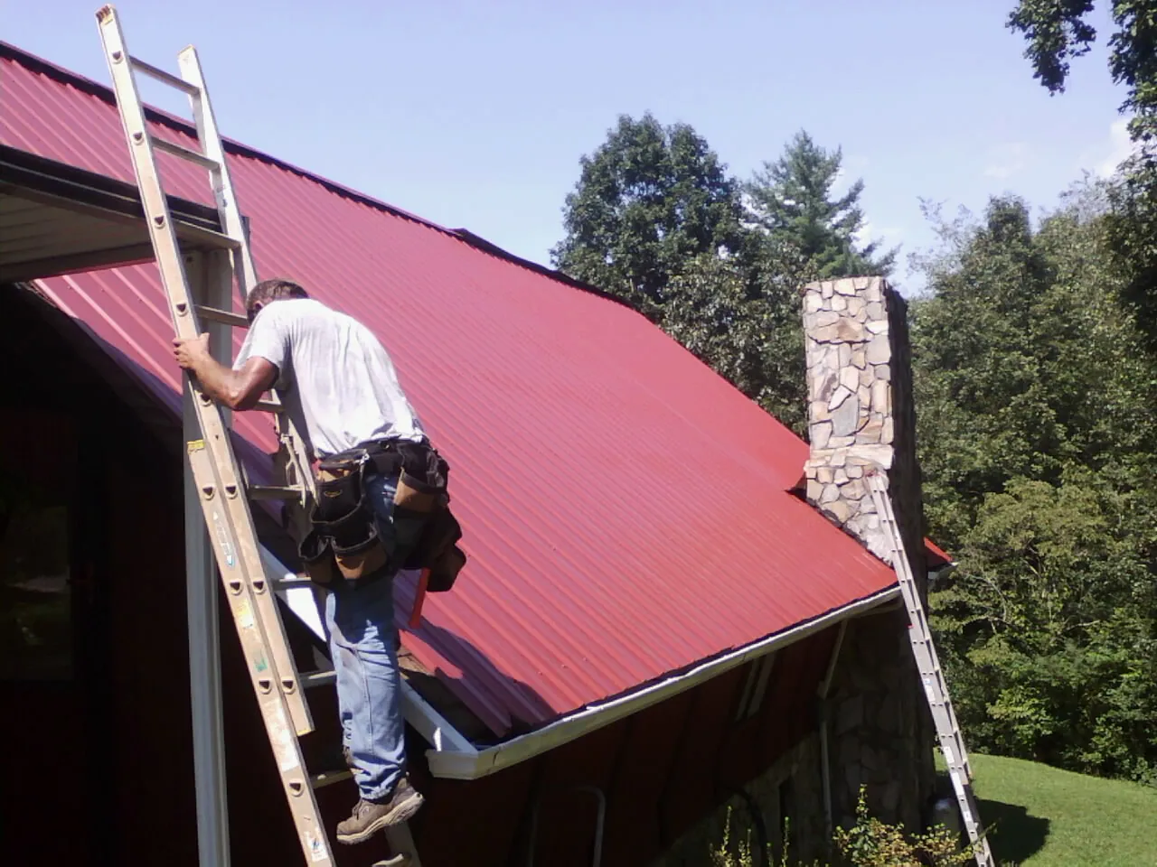 Roofer on a ladder beside a red metal roof and stone chimney — Soffit & Fascia Repair by Roofing Brothers in Bedford
